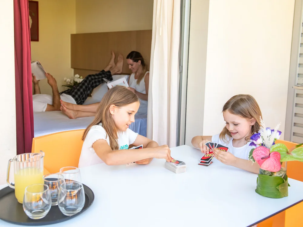 Bibione Palace Hotel · La tua casa al mare a 4 stelle S Due bambine giocano a carte a un tavolo con un vaso di fiori e una famiglia sul letto