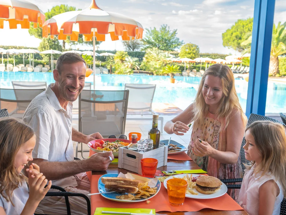Bibione Palace Hotel · La tua casa al mare a 4 stelle S Famiglia che mangia all'aperto vicino a una piscina con ombrelloni arancioni