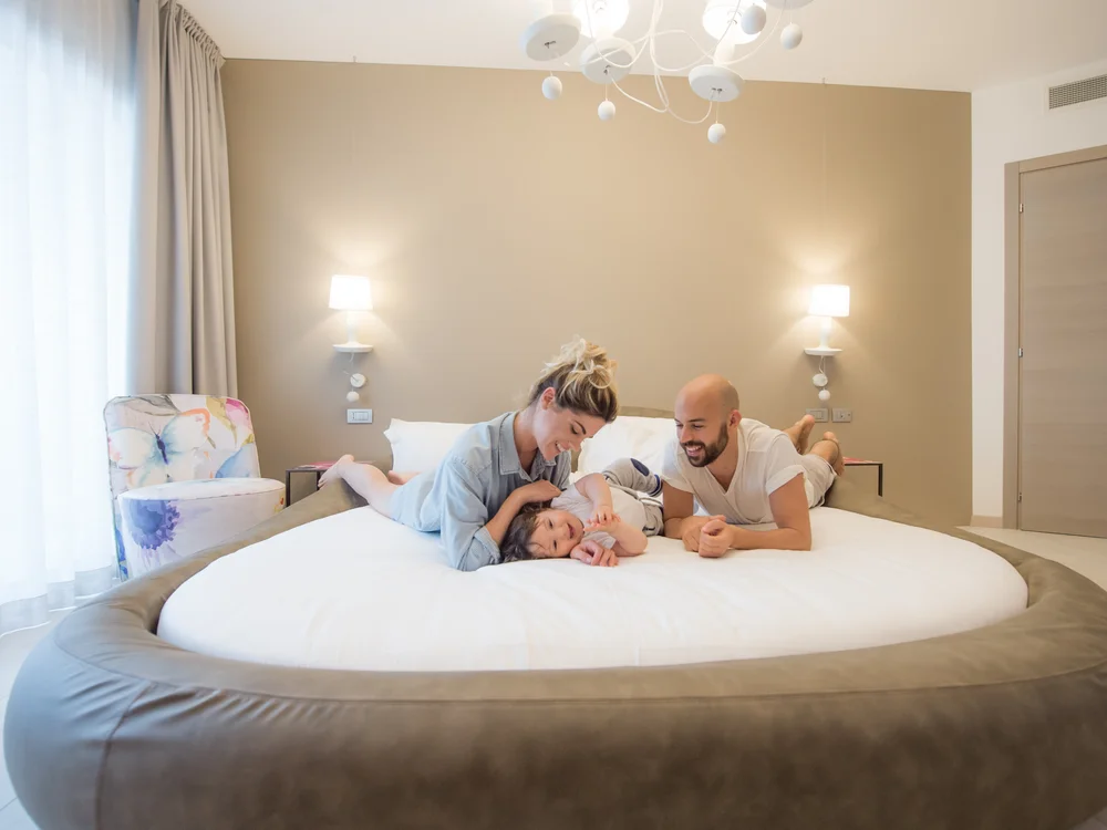 Bibione Palace Hotel · La tua casa al mare a 4 stelle S Famiglia felice sdraiata su un grande letto rotondo in una stanza luminosa