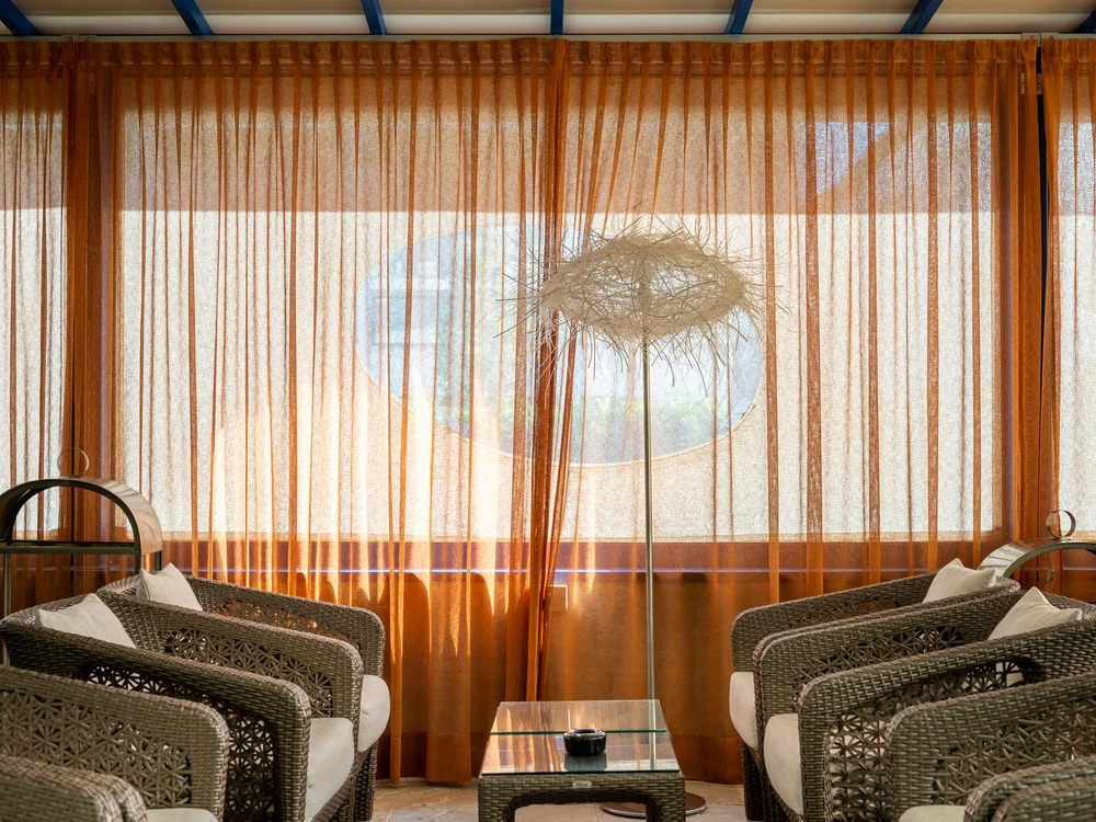 Bibione Palace Hotel · La tua casa al mare a 4 stelle S Salotto con poltrone in vimini, tavolino e tende arancioni trasparenti