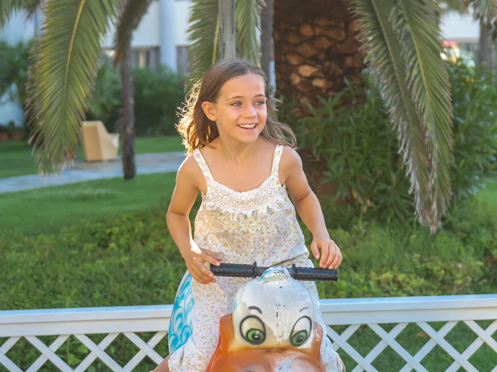 Bibione Palace Hotel · La tua casa al mare a 4 stelle S Bambina sorridente che gioca su un'altalena a forma di uccello arancione in giardino