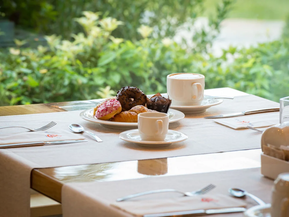 Bibione Palace Hotel · La tua casa al mare a 4 stelle S Colazione con caffè e dolci su un tavolo apparecchiato vicino a una finestra verde