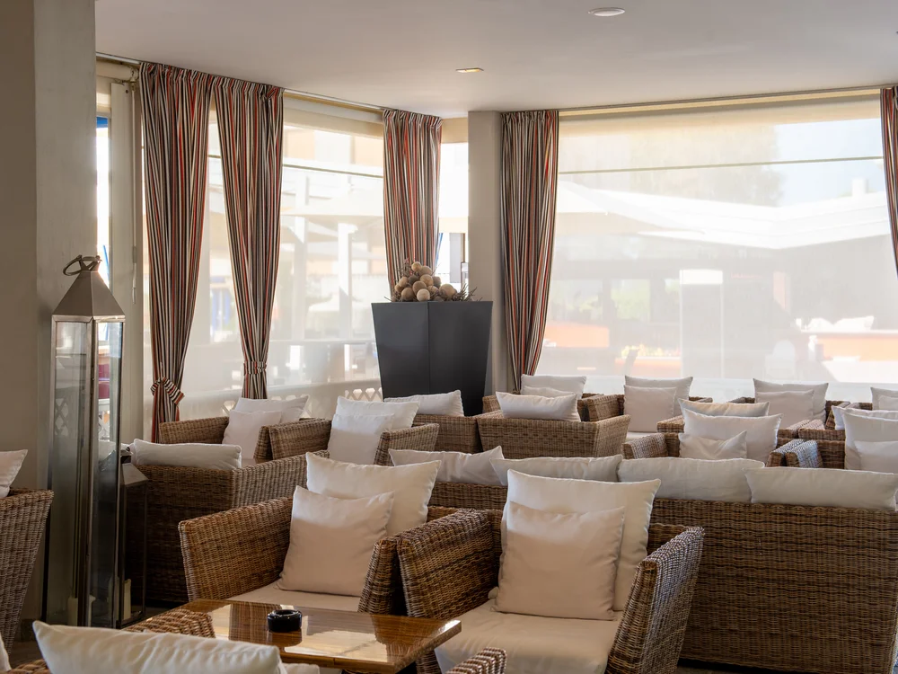 Bibione Palace Hotel · La tua casa al mare a 4 stelle S Area lounge con poltrone in vimini e cuscini bianchi vicino a finestre con tende