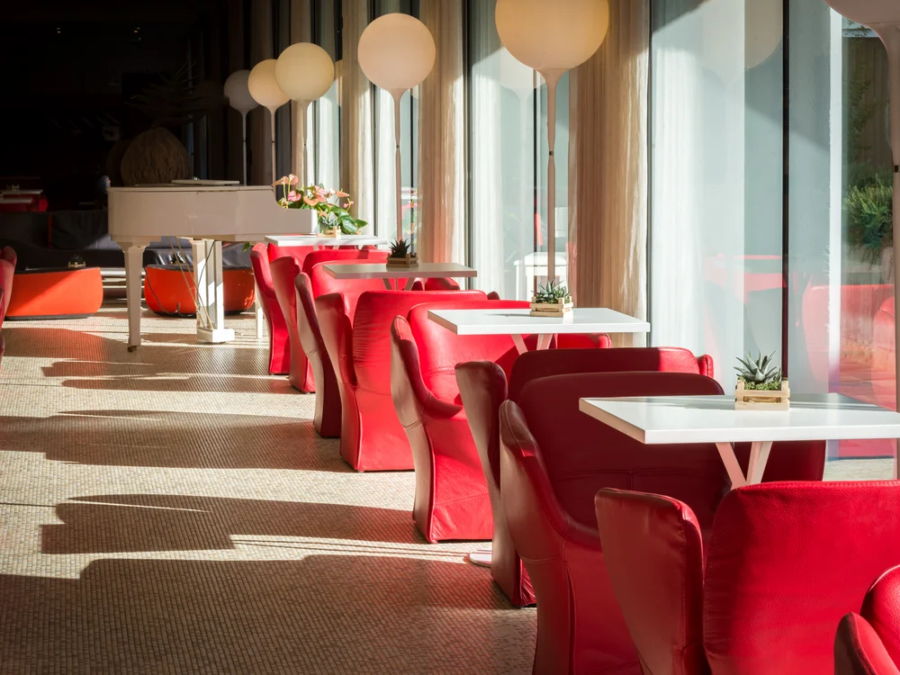Bibione Palace Hotel · La tua casa al mare a 4 stelle S Sala luminosa con poltrone rosse, tavoli bianchi e pianoforte bianco