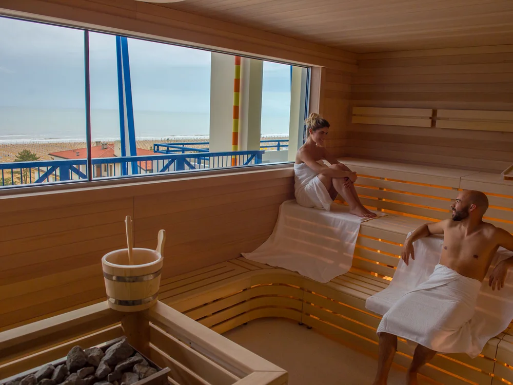 Bibione Palace Hotel · La tua casa al mare a 4 stelle S Due persone rilassate in sauna con vista sulla spiaggia attraverso la finestra