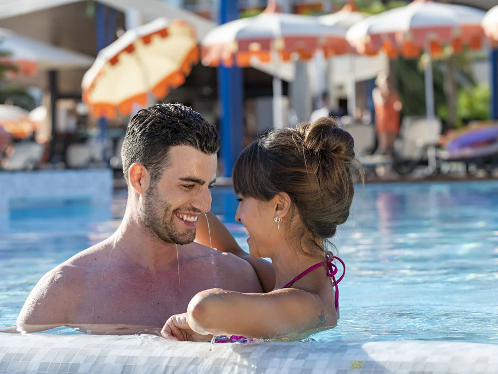 Bibione Palace Hotel · La tua casa al mare a 4 stelle S Coppia sorridente che si abbraccia in piscina con ombrelloni sullo sfondo