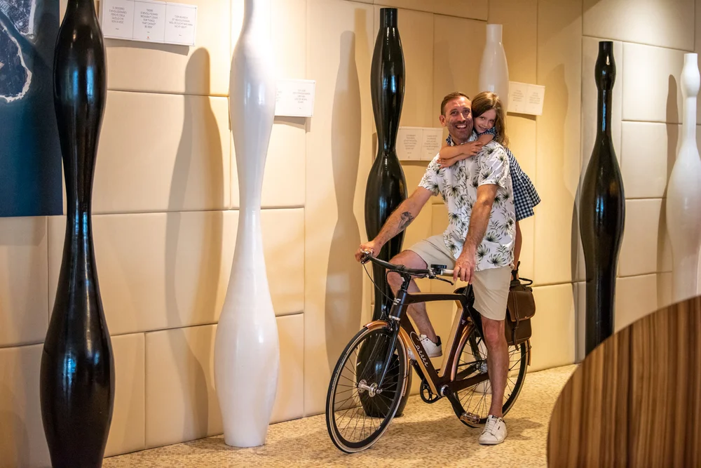 Uomo con bambina sulla schiena su bicicletta in corridoio con sculture moderne