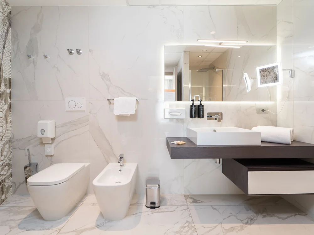 Bibione Palace Hotel · La tua casa al mare a 4 stelle S Bagno moderno con sanitari bianchi e lavabo con specchio illuminato