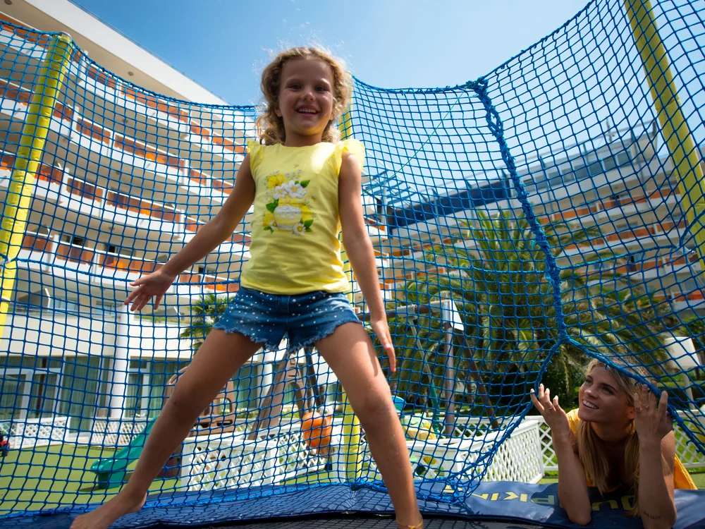 Bibione Palace Hotel · La tua casa al mare a 4 stelle S Bambina che salta felice su un trampolino con rete blu in un parco giochi all'aperto