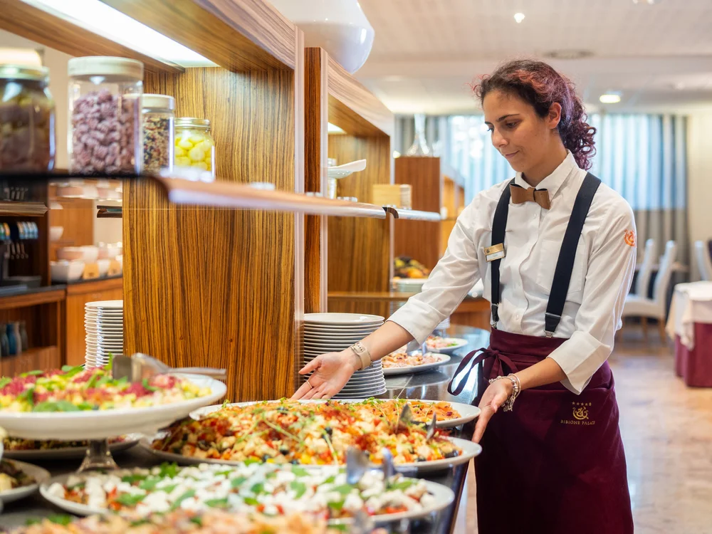 Bibione Palace Hotel · La tua casa al mare a 4 stelle S Cameriera che sistema piatti di cibo su un buffet in un ristorante