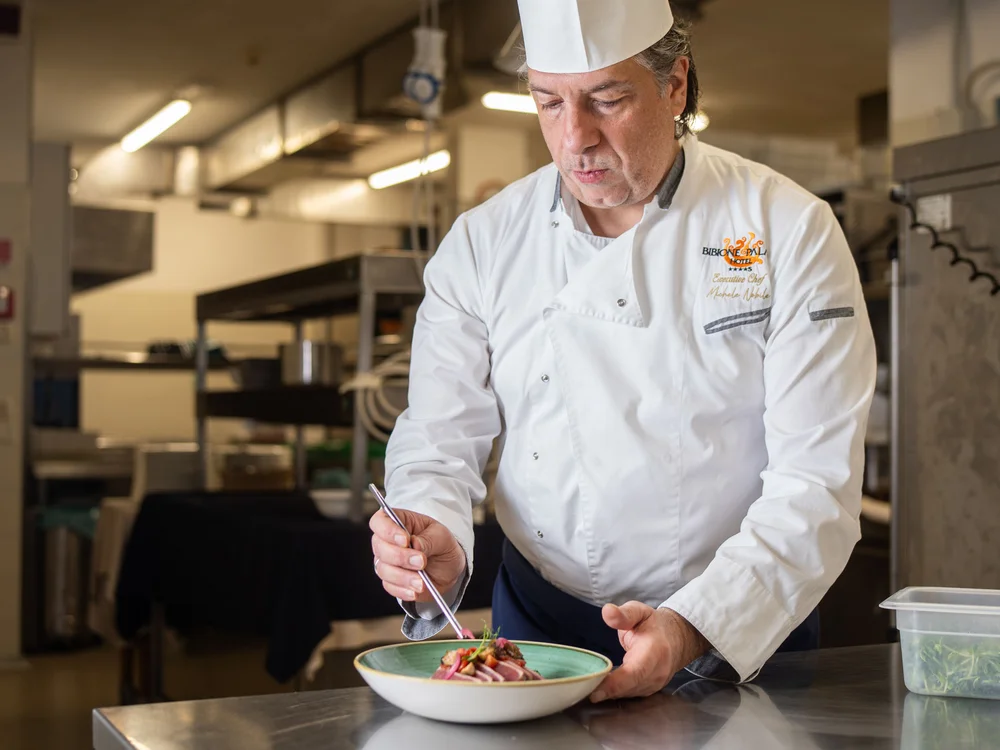 Bibione Palace Hotel · La tua casa al mare a 4 stelle S Chef in uniforme bianca che decora un piatto in cucina professionale